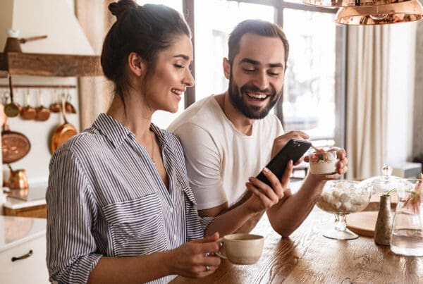 Read Our Reviews - Young Couple Smiles as They Use a Smartphone in Their Bright Kitchen, Enjoying Coffee